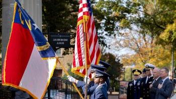 Uniformed ROTC members holding North Carolina and American flags as part of a Veterans Day ceremony at UNC-Chapel Hill outside of Memorial Hall. In the background are men holding their hands across their chest and others in military uniform saluting.