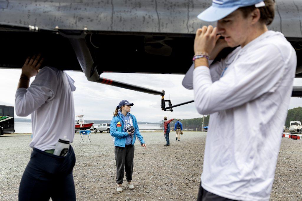 Karen Jordan standing on beach wearing a blue jacket and baseball cap as she watches the rowing team prepare.